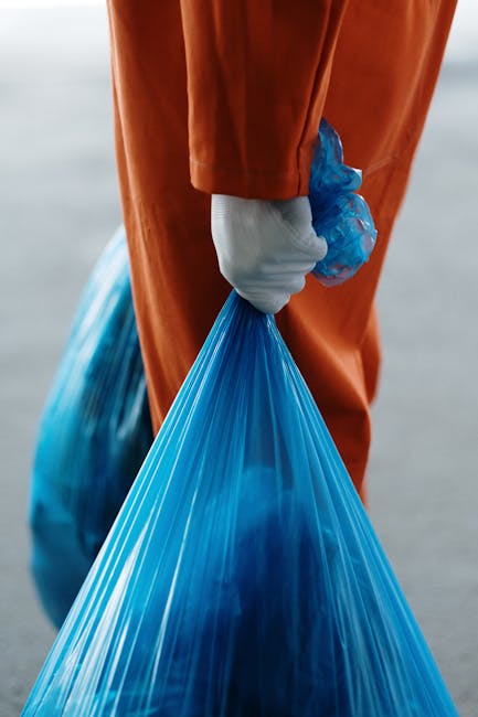 A person wearing orange work overalls and white sneakers standing on a grey carpeted floor, holding two large blue plastic trash bags filled with waste in each hand. The background features a plain li