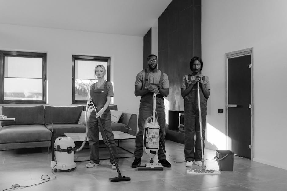 A team of three professional cleaners from Cleaners Hounslow are performing surface cleaning and deep cleaning in a modern living room. The room features a large, L-shaped sofa with a dark fabric upholstery, positioned against a wall with two large windows that let in natural light. The flooring is light-colored wooden, visibly clean and shiny. The middle cleaner, a man, stands confidently with a handheld vacuum cleaner in front of a sleek, dark wood panel on the wall, while the two female cleaners on either side hold cleaning tools; one has a vacuum hose connected to a separate canister, and the other is holding a mop with a bucket nearby. The room's decor is minimalist, with neutral tones and contemporary furnishings, highlighting the cleanliness and hygiene achieved through thorough surface cleaning. Cleaners Hounslow consistently uses high-quality products and equipment to ensure effective sanitisation and maintenance, aligned with their domestic cleaning services. The setting emphasizes the importance of professional cleaning for maintaining a hygienic and inviting home environment.