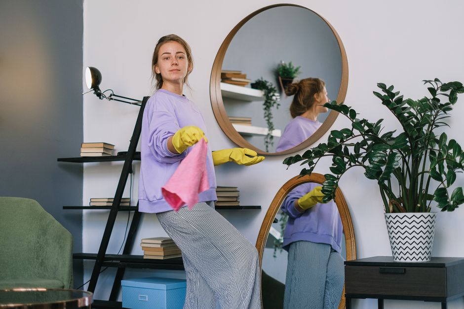 A team of three professional cleaners from Cleaners Hounslow are performing surface cleaning and deep cleaning in a modern living room. The room features a large, L-shaped sofa with a dark fabric upholstery, positioned against a wall with two large windows that let in natural light. The flooring is light-colored wooden, visibly clean and shiny. The middle cleaner, a man, stands confidently with a handheld vacuum cleaner in front of a sleek, dark wood panel on the wall, while the two female cleaners on either side hold cleaning tools; one has a vacuum hose connected to a separate canister, and the other is holding a mop with a bucket nearby. The room's decor is minimalist, with neutral tones and contemporary furnishings, highlighting the cleanliness and hygiene achieved through thorough surface cleaning. Cleaners Hounslow consistently uses high-quality products and equipment to ensure effective sanitisation and maintenance, aligned with their domestic cleaning services. The setting emphasizes the importance of professional cleaning for maintaining a hygienic and inviting home environment.