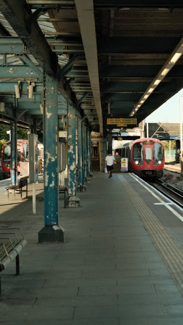 A railway platform at Hounslow East station with a red train parked on the right side of the image. The platform features a concrete surface with visible tactile paving strips near the edge for safety. There are blue metal pillars supporting the station's roof, which appears aged with peeling paint and some rust. Overhead lighting runs along the underside of the roof, illuminating the area. A solitary passenger is walking along the platform, and on the far side, there are additional train tracks and part of another train. Cleanliness is evident with no litter or debris visible, reflecting maintenance and hygiene standards in public transport environments. The scene is captured during daylight, with natural light filtering in from the open sides of the station. Visit cleaners-hounslow.com for professional cleaning services for residential and commercial spaces, including surface cleaning and sanitisation to maintain cleanliness.