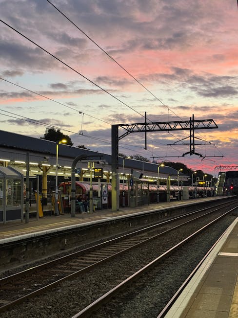 A view of Hounslow East railway station during sunset with multiple train tracks running parallel along the platform. The platform is protected by a glass shelter with metal framing, and it is illuminated by bright yellow lighting fixtures. Several modern trains are parked at the station, featuring smooth metallic surfaces and tinted windows. Overhead power lines run above the tracks, supported by tall metal gantries, with faint electrical wires spanning across the sky. The backdrop shows a colorful evening sky with shades of pink, orange, and purple, and some silhouettes of trees in the distance. This scene exemplifies a typical train station setting, emphasizing clean, maintained surfaces and organized layout, which is essential for safe and efficient transit. Cleaners Hounslow provides professional cleaning services ensuring platforms and stations remain hygienic, tidy, and ready for daily commuters.