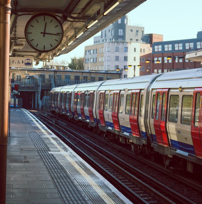 A railway platform at Hounslow East station with a red train parked on the right side of the image. The platform features a concrete surface with visible tactile paving strips near the edge for safety. There are blue metal pillars supporting the station's roof, which appears aged with peeling paint and some rust. Overhead lighting runs along the underside of the roof, illuminating the area. A solitary passenger is walking along the platform, and on the far side, there are additional train tracks and part of another train. Cleanliness is evident with no litter or debris visible, reflecting maintenance and hygiene standards in public transport environments. The scene is captured during daylight, with natural light filtering in from the open sides of the station. Visit cleaners-hounslow.com for professional cleaning services for residential and commercial spaces, including surface cleaning and sanitisation to maintain cleanliness.