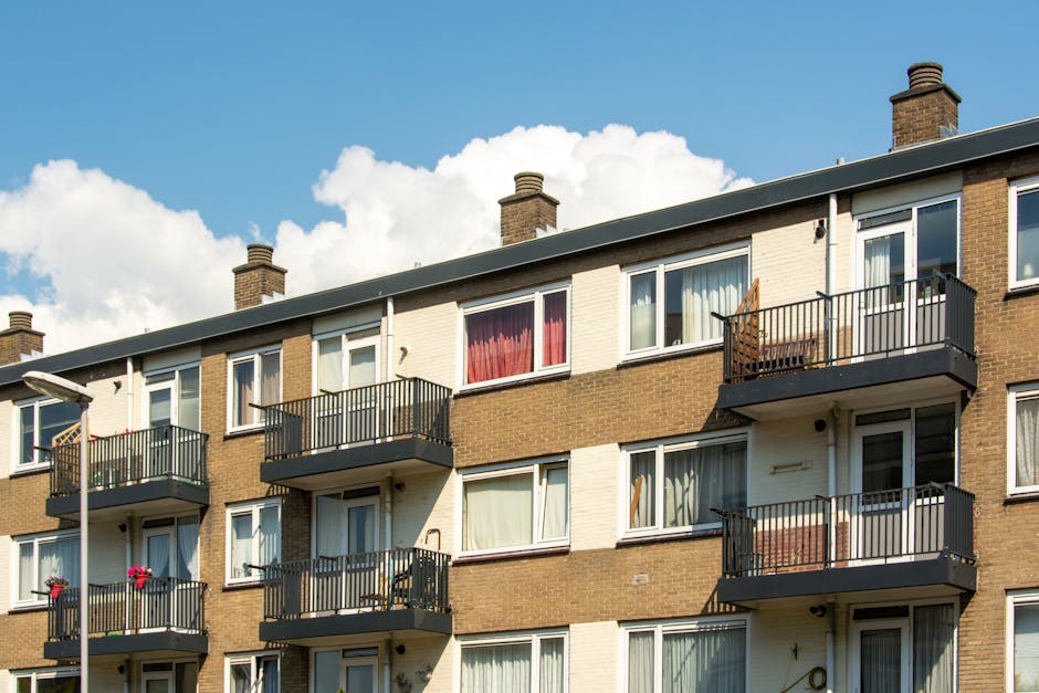 Photograph of a multi-story residential apartment building with a brown brick facade and white sections. Each apartment has a balcony with black metal railings, some with visible outdoor furniture and
