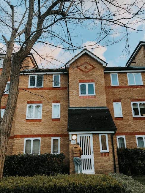 Photograph of a multi-story residential apartment building with a brown brick facade and white sections. Each apartment has a balcony with black metal railings, some with visible outdoor furniture and