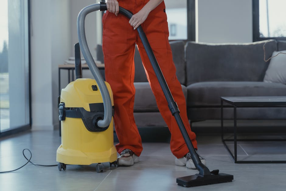 A professional cleaner wearing orange work trousers and grey shoes is using a yellow vacuum cleaner with a black hose to perform surface cleaning on a tiled floor in a modern living room. The vacuum cleaner is placed on the floor, and the worker is holding the hose and nozzle to clean the tile surface, which appears clean and dry. In the background, there is a dark grey sofa with cushions and a glass side table, with natural light streaming in through large windows, creating a bright and airy environment. This image illustrates deep cleaning services provided by Cleaners Hounslow, focusing on maintaining hygiene and cleanliness in residential spaces, specifically on floor surface care and routine maintenance.