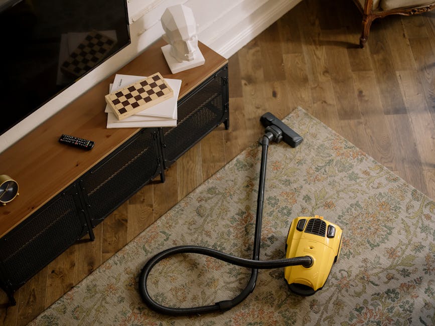 A yellow canister vacuum cleaner with a black hose and metal wand attachment resting on a patterned area rug in a living room. The room has wooden flooring, and a black sideboard with a wooden top is visible, holding a white abstract bust, a small plant, a remote control, and a chessboard. The lighting appears natural and highlights the clean, polished surfaces and tidy space, indicative of professional carpet cleaning services provided by Cleaners Hounslow in the context of surface cleaning and hygiene maintenance for residential interiors.