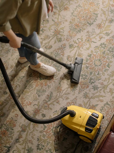 A professional cleaner wearing orange work trousers and grey shoes is using a yellow vacuum cleaner with a black hose to perform surface cleaning on a tiled floor in a modern living room. The vacuum cleaner is placed on the floor, and the worker is holding the hose and nozzle to clean the tile surface, which appears clean and dry. In the background, there is a dark grey sofa with cushions and a glass side table, with natural light streaming in through large windows, creating a bright and airy environment. This image illustrates deep cleaning services provided by Cleaners Hounslow, focusing on maintaining hygiene and cleanliness in residential spaces, specifically on floor surface care and routine maintenance.