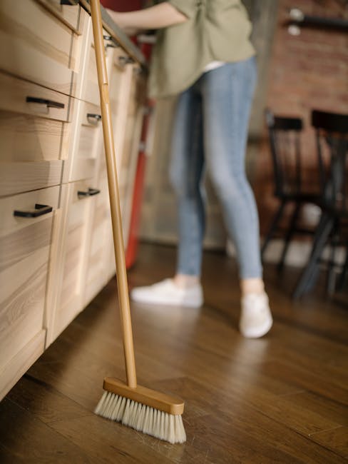 Close-up view of a wooden-handled broom with white bristles resting on a polished wooden floor in a kitchen or dining area. In the background, a person wearing light blue jeans and white sneakers is standing near a light wood kitchen cabinet, with a countertop and various kitchen items partially visible. The room is well-lit, showcasing the clean and tidy environment. The image emphasizes surface cleaning and maintenance typical of a domestic cleaning service provided by Cleaners Hounslow for period homes in Osterley Park.