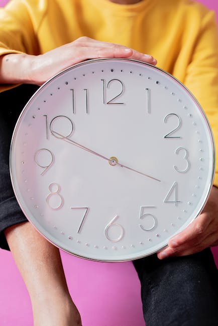 A person wearing a yellow long-sleeve shirt is holding a large, white round wall clock with a minimalist design, featuring embossed numbers and thin gray hands showing a time of approximately 10:50. The background is a solid pink color, and the person is seated with their hand resting lightly on top of the clock. The clock's surface appears smooth and clean, emphasizing its role in surface cleaning and time management, relevant to same-day domestic cleaning services offered by Cleaners Hounslow, particularly in the context of avoiding last-minute booking delays in Hounslow.